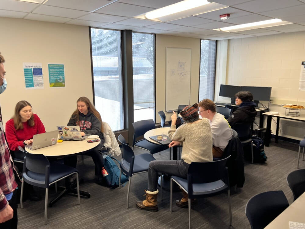 Students sitting in groups at tables in a bright classroom, working on laptops and engaging in discussion, with large windows, posters on the wall, and snacks on a side table